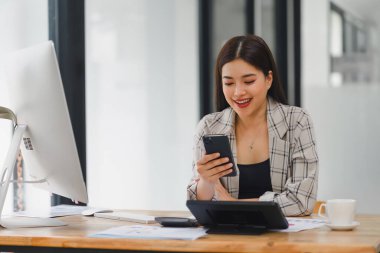 Ofiste akıllı telefon kullanan Asyalı bir iş kadını. Masasında masaüstü bilgisayarı ve mali raporları var..