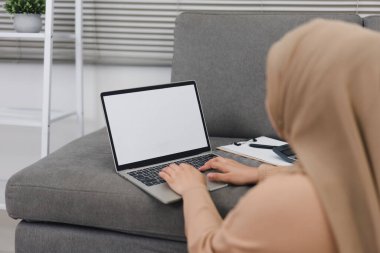 Başörtüsü takan Müslüman kadın, koltukta otururken beyaz ekran dizüstü bilgisayar kullanarak satış performans grafiklerini analiz ediyor.