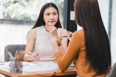 İki genç Asyalı iş kadını bir yatırım projesi ve planlama stratejisini tartışıyor ofiste dizüstü bilgisayarla çalışırken sohbet ediyorlar.