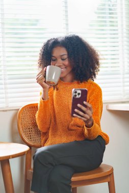 Turuncu kazaklı, güzel, Afro-Amerikan kadın. Kahvesini içiyor, akıllı telefon kullanıyor. Parlak, modern bir ofis odasında dinleniyor.