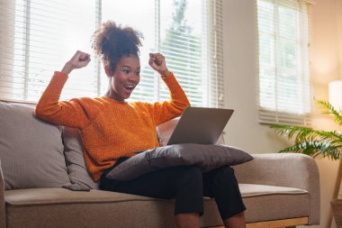 Genç siyahi kadın, rahat oturma odasında dizüstü bilgisayarla çalışırken başarıyı kutluyor.