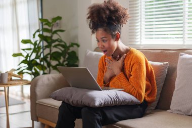 Heyecanlı Afrikalı Amerikalı genç bir kadın evinde, rahat bir oturma odasında çevrimiçi aktivitelere katılıyor.
