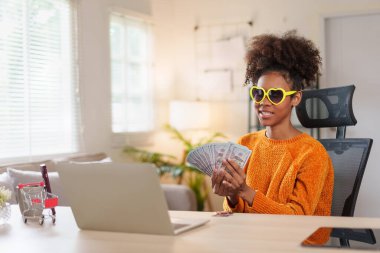 Kalp şeklinde güneş gözlüğü takan neşeli bir kadın, elinde dolarların hayranı, kameraya gülümseyen, finansal başarı ve mutluluğu ifade eden.