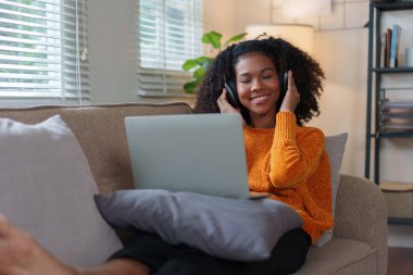 Gülümseyen Afrikalı Amerikalı kadın rahat bir kanepede dinleniyor, kulaklık takıyor, dizüstü bilgisayar kullanıyor, rahat bir evin tadını çıkarıyor..