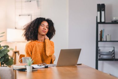 Evde dizüstü bilgisayarla çalışırken boynuna masaj yapan Afro-Amerikalı kadın rahatsızlığını ya da gerginliğini belirtiyor.