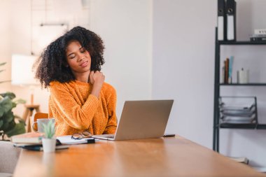 Evde dizüstü bilgisayarla çalışırken boynuna masaj yapan Afro-Amerikalı kadın rahatsızlığını ya da gerginliğini belirtiyor.