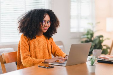Gözlüklü mutlu Afro-Amerikalı kadın evde dizüstü bilgisayarla çalışıyor.