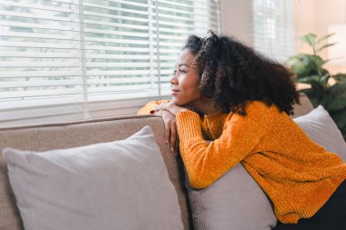 Düşünceli Afrikalı Amerikalı kadın, kanepede dinleniyor, sakin bir ifadeyle pencereden dışarı bakıyor.