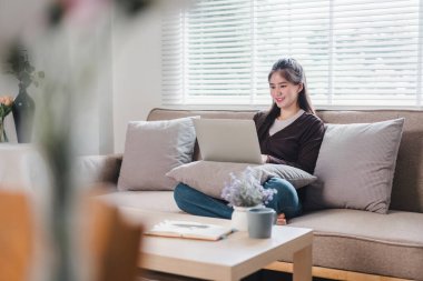 Gülümseyen genç Asyalı kadın koltukta otururken dizüstü bilgisayar kullanıyor, evinde rahat ve rahat bir anın tadını çıkarıyor.