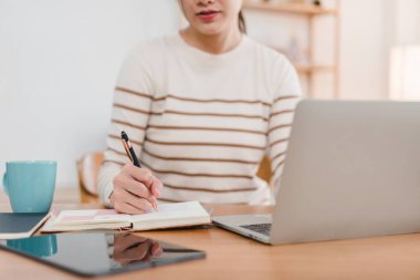Asyalı iş kadını laptopuyla ev ofisinde çalışırken not alıyor.