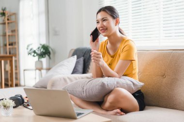 Sarı tişörtlü bir kadın elinde akıllı bir telefonla dizüstü bilgisayarıyla çalışır, birden fazla iş yapmaya ve bağlantıda kalmaya odaklanır..