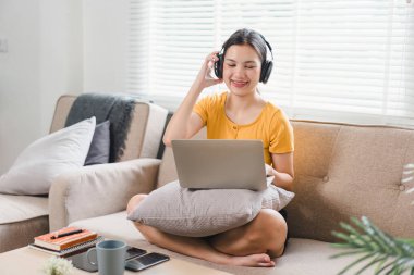 Gülümseyen Asyalı kadın rahat bir koltukta dinleniyor, kulaklık takıyor, dizüstü bilgisayar kullanıyor, rahat bir evin tadını çıkarıyor.