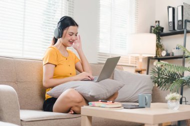 Gülümseyen Asyalı kadın rahat bir koltukta dinleniyor, kulaklık takıyor, dizüstü bilgisayar kullanıyor, rahat bir evin tadını çıkarıyor.