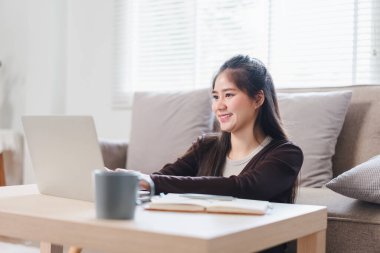Gülümseyen genç Asyalı kadın koltukta otururken dizüstü bilgisayar kullanıyor, evinde rahat ve rahat bir anın tadını çıkarıyor.
