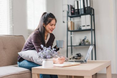 Bir kadın rahat bir şekilde kanepede oturuyor, bir fincan kahvenin tadını çıkarırken dizüstü bilgisayarıyla çalışıyor. Sıcak atmosfer, doğal ışık ve masadaki küçük bitki ile gelişerek üretken ve rahat bir envir oluşturur.