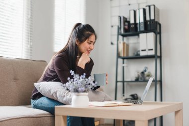 Bir kadın, dizüstü bilgisayarıyla uğraşırken rahatça kanepede oturup bir fincan kahvenin tadını çıkarıyor. Sıcak atmosfer, doğal ışık ve şık iç mekan ile gelişir..