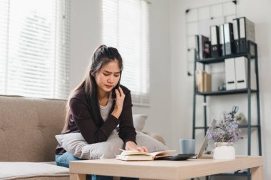 Kanepede oturan bir kadın, telefonla konuşurken endişeli görünüyor, rahat bir oturma odası atmosferiyle çevrili. Kitap okuyor ve yakınlarda dizüstü bilgisayarı var. Odaklanma ve düşünme hissi yaratıyor..