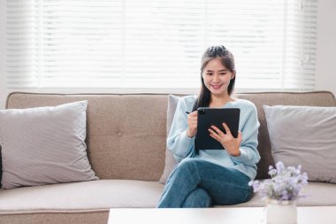 Genç bir kadın kanepede rahatça oturuyor, gülümserken tablet kullanıyor. Rahat oturma odasında doğal ışık ve dekoratif yastıklar var. Sıcak atmosfer yaratıyorlar..
