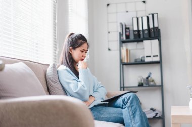 Genç bir kadın düşünceli bir şekilde kanepede oturur, elinde tabletle rahat, aydınlık bir oturma odasında. Dalgın görünüyor. Etrafı sakin bir atmosferle çevrili..