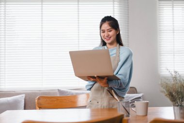 Genç bir kadın, parlak ve rahat bir odada dizüstü bilgisayar kullanırken gülümsüyor. Meşgul ve mutlu görünüyor, modern çalışma alanını doğal ışıkla gösteriyor..