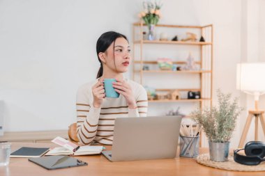 Masasında oturan genç bir kadın, elinde mavi kupayla dikkatlice dizüstü bilgisayarından uzağa bakıyor. Çalışma alanı parlak ve düzenlidir, bitkiler ve dekoratif malzemelerle, sakin bir atmosfer yaratır..