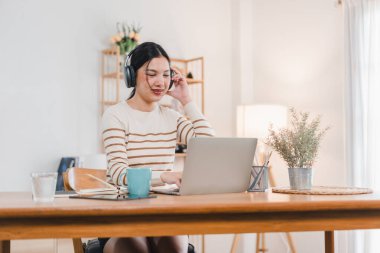 Kulaklık takan genç bir kadın, parlak ve modern çalışma alanındaki ahşap masada otururken dizüstü bilgisayarına odaklanıyor. Bitkiler ve rahat atmosferle çevrili, meşgul ve üretken görünüyor..