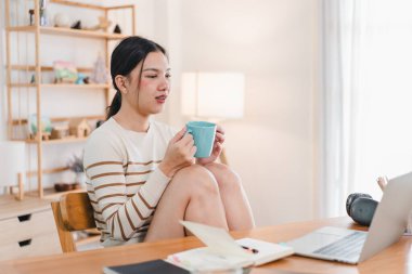 Genç bir kadın, rahat ev ortamlarıyla çevrili ahşap masada otururken sıcak içkiden zevk alır. Rahatlamış ve laptopuna odaklanmış görünüyor. Huzurlu bir atmosfer yaratıyor..