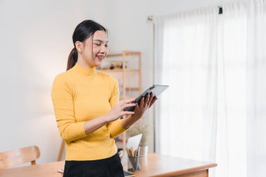 Sarı kazaklı genç bir kadın, modern çalışma alanında tablet kullanırken gülümsüyor. Neşeli tavırları çevreye olumlu bir hava katıyor..