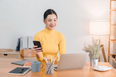 Sarı boğazlı kazaklı genç bir kadın ahşap masada akıllı telefonunu ve laptopunu kullanırken gülümsüyor. Sıcak atmosfer yaratan konforlu çalışma alanı bitkiler, defterler ve kupalar..