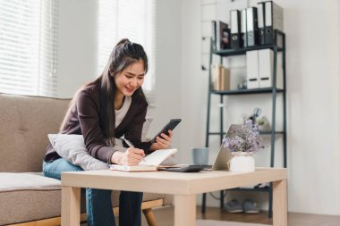 Asian woman using laptop, reviewing financial documents, managing bills and budgeting in cozy home clipart