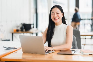 Kendine güvenen bir kadın, modern ofiste dizüstü bilgisayarla çalışırken gülümser..