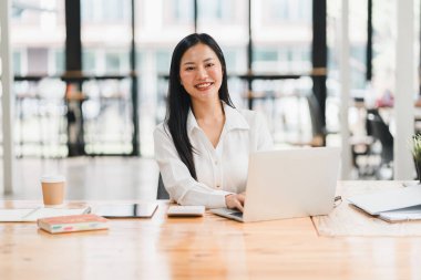 Modern ofis alanında dizüstü bilgisayarda çalışan etkili bir kadın, mutlu bir şekilde gülümsüyor..