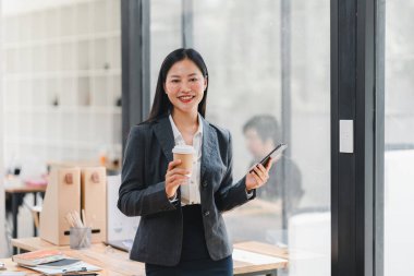 Takım elbiseli bir kadın modern ofiste kahve ve telefon tutuyor..