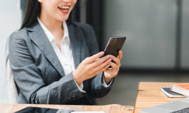 Profesyonel bir kadın masasında akıllı telefon kullanırken gülümser. Pozitif çalışma ortamı sunar..