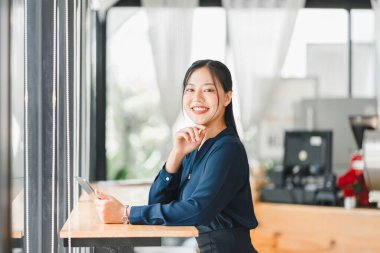 Kendine güvenen bir kadın kafede oturmuş elinde tabletle gülümsüyor..