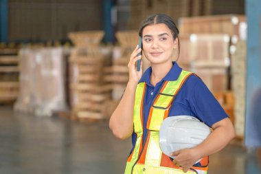 Depoda cep telefonuyla duran genç bayan depo çalışanı kasketli gülümseyen kadın fabrika deposunda lojistik sektöründe çalışan işçi.