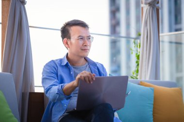 Akıllı telefon ve dizüstü bilgisayar kullanan Asyalı bir adam evdeki koltukta oturuyor, terasta dizüstü bilgisayarla çalışan bir adam.