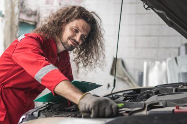 Kaputu açık, kontrol listesi olan genç beyaz araba tamircisi, garajda modern arabayı kontrol eden teknisyen, araba tamiri ve bakım konsepti.