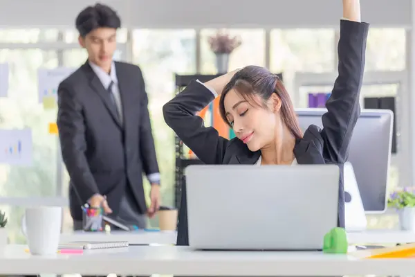Asian woman doing stretch exercise stretching her arms in work time, Female officer feeling tired and fatigue stretching to relax with blurred colleagues in modern office