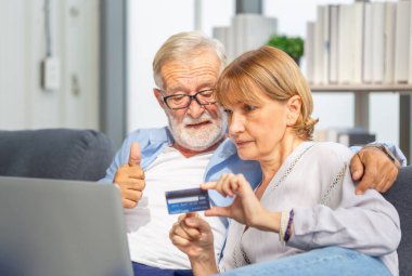 Bilgisayarlı yaşlı çift evde online alışveriş yapıyor. Yakışıklı yaşlı adam başparmaklarını kaldırıyor ve elinde kredi kartı tutan kadın