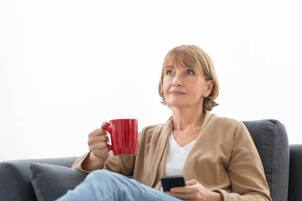 Portrait of Relaxing woman with cups of coffee on cozy sofa in the living room, Mature woman using mobile smartphone during a coffee break
