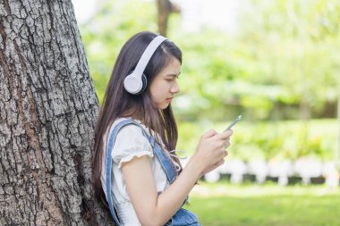 Ağaç gövdesinde oturan genç bir kız parkta kulaklıkla müzik dinliyor, genç Asyalı bir kadın müzikten hoşlanıyor.