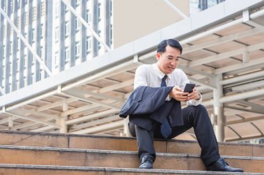 Asyalı işadamı merdivenlerde oturuyor ve ofisin dışındaki cep telefonunda çalışmalarını kontrol ediyor.