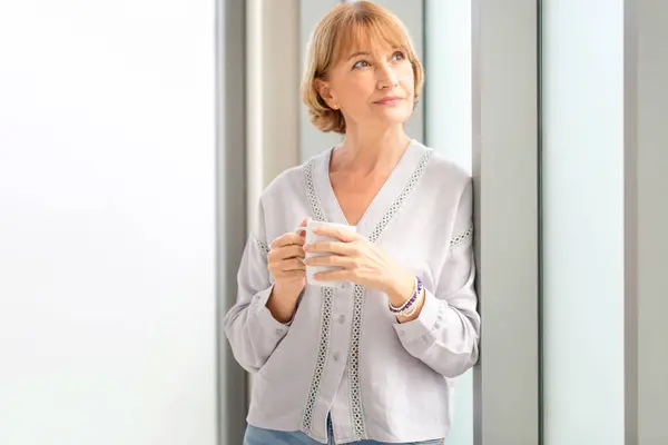 Relaxing woman standing near window with cups of coffee, woman inside new home during a coffee break