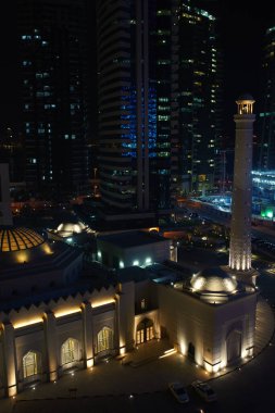 DOHA, QATAR - SEPTEMBER 16, 2019: Ibrahim Khalil Al-Darwish Mosque surrounded by skyscrappers on the hot night clipart