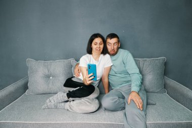 Millennial couple with disgust face expression because aversion reaction looking at smartphone screen sitting on sofa at home. Human emotions concept