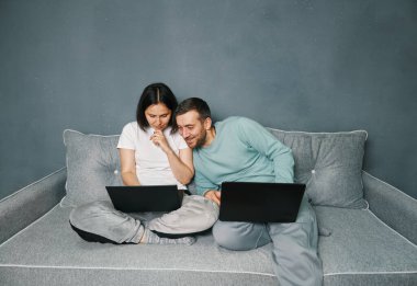 Young smiling couple relaxing on sofa with their laptops surfing the Internet, enjoying work from home.
