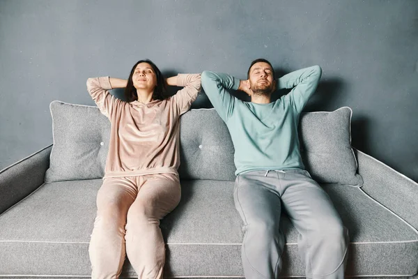 Couple relaxing on a sofa at home stretching with hands behind head. Lazy weekend, rest concept