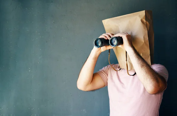 Man with a paper bag on head looking through binoculars to copy space over gray background