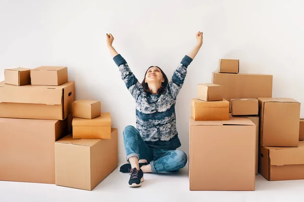 Happy young woman celebrate moving to new home with raised hands sitting on floor surrounded by cardboard boxes. Real estate, relocation concept
